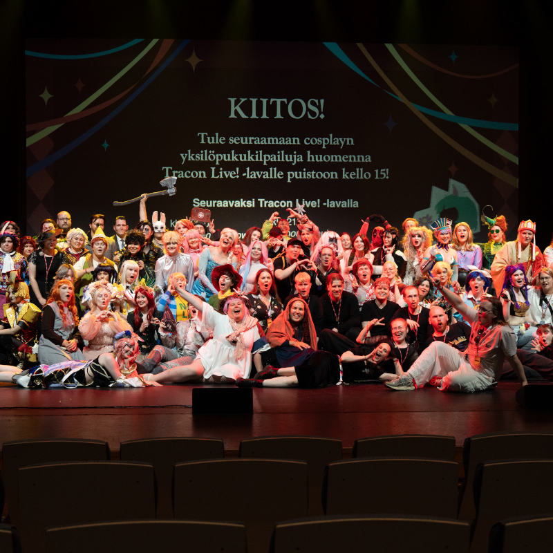 A photo of a merry group of cosplayers and cosplay competition organisers posing for the camera on the main stage of Tracon. In the background, the screen says 'Kiitos', which is 'thank you' in Finnish. The photo is taken by Emilmeemu.