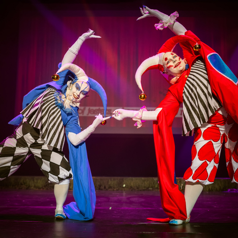 Two cosplayers as Zorn and Thorn from Final Fantasy IX, red and blue jester-like characters, leaning towards each other and forming a circle with their hands. The photo is taken by Nyymix.
